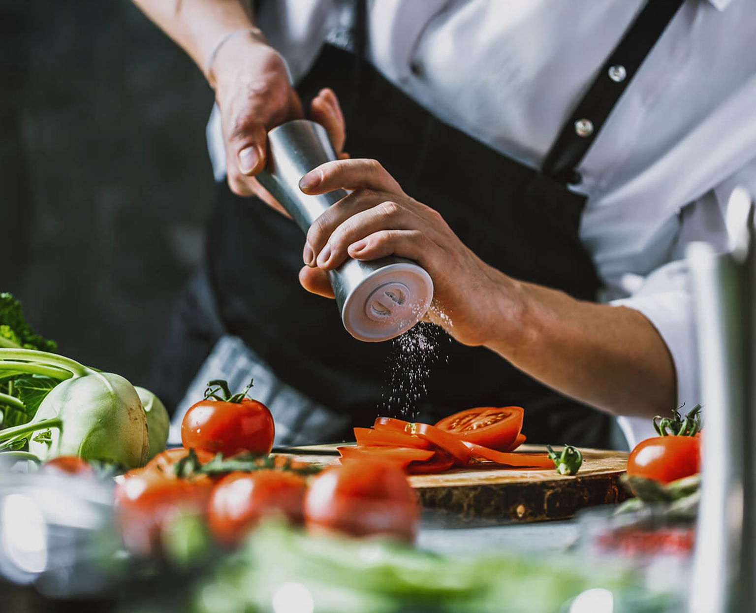A person wearing a white shirt and black apron grinds fresh pepper onto sliced tomatoes on a wooden cutting board, surrounded by tomatoes and green vegetables.