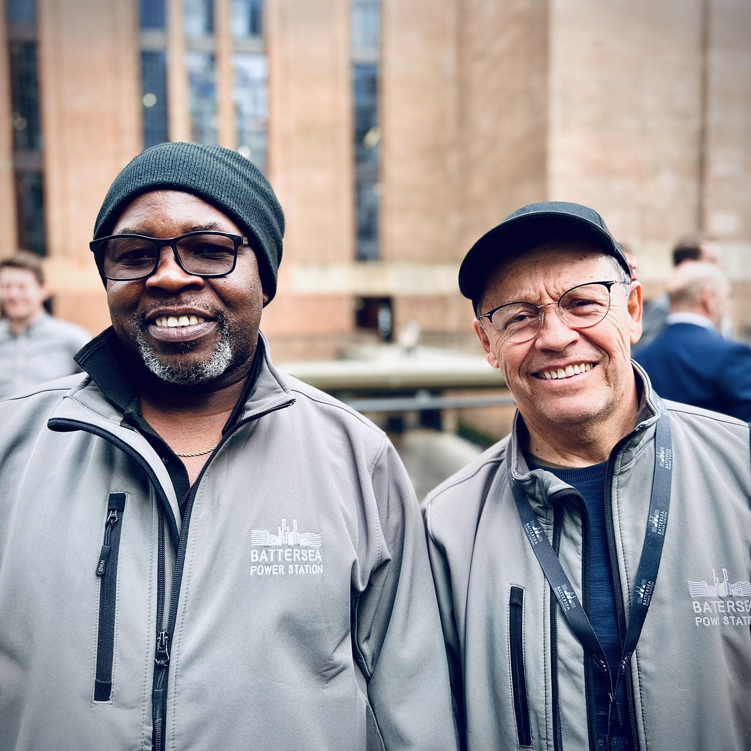 Two smiling men wearing glasses, hats, and matching grey Battersea Power Station jackets stand side by side outdoors in front of a large brick building. Other people are blurred in the background.