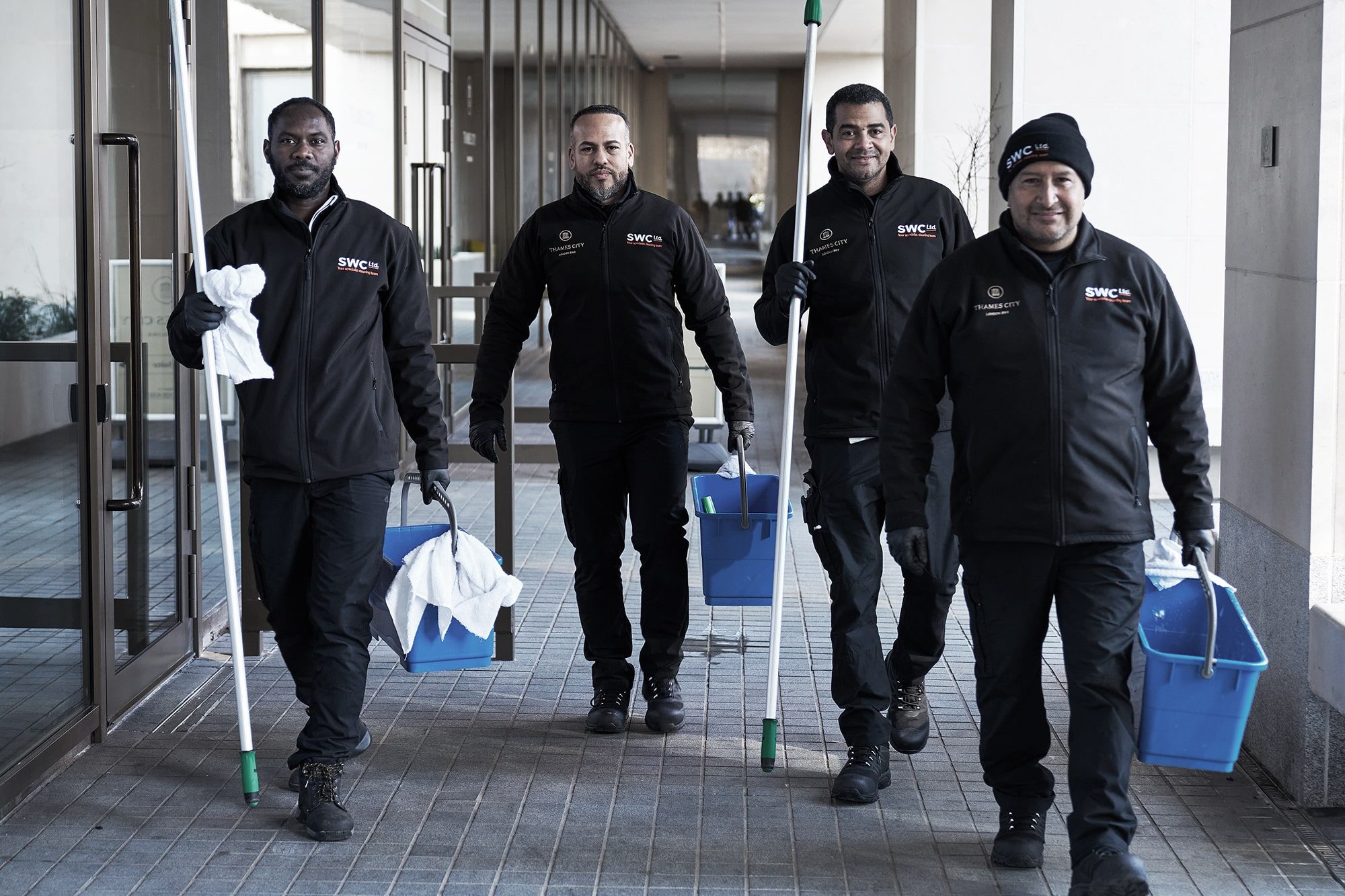 Four janitors wearing black uniforms walk outside a building. Two carry mops and blue buckets, another has a white towel, and all appear to be heading to work. The group looks focused and professional.