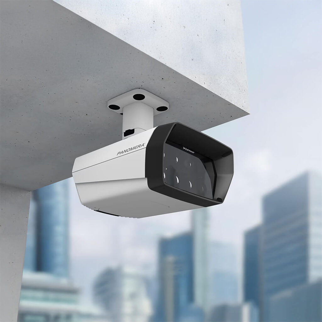 A modern security camera mounted under a concrete overhang, with a cityscape of tall, glass buildings blurred in the background.