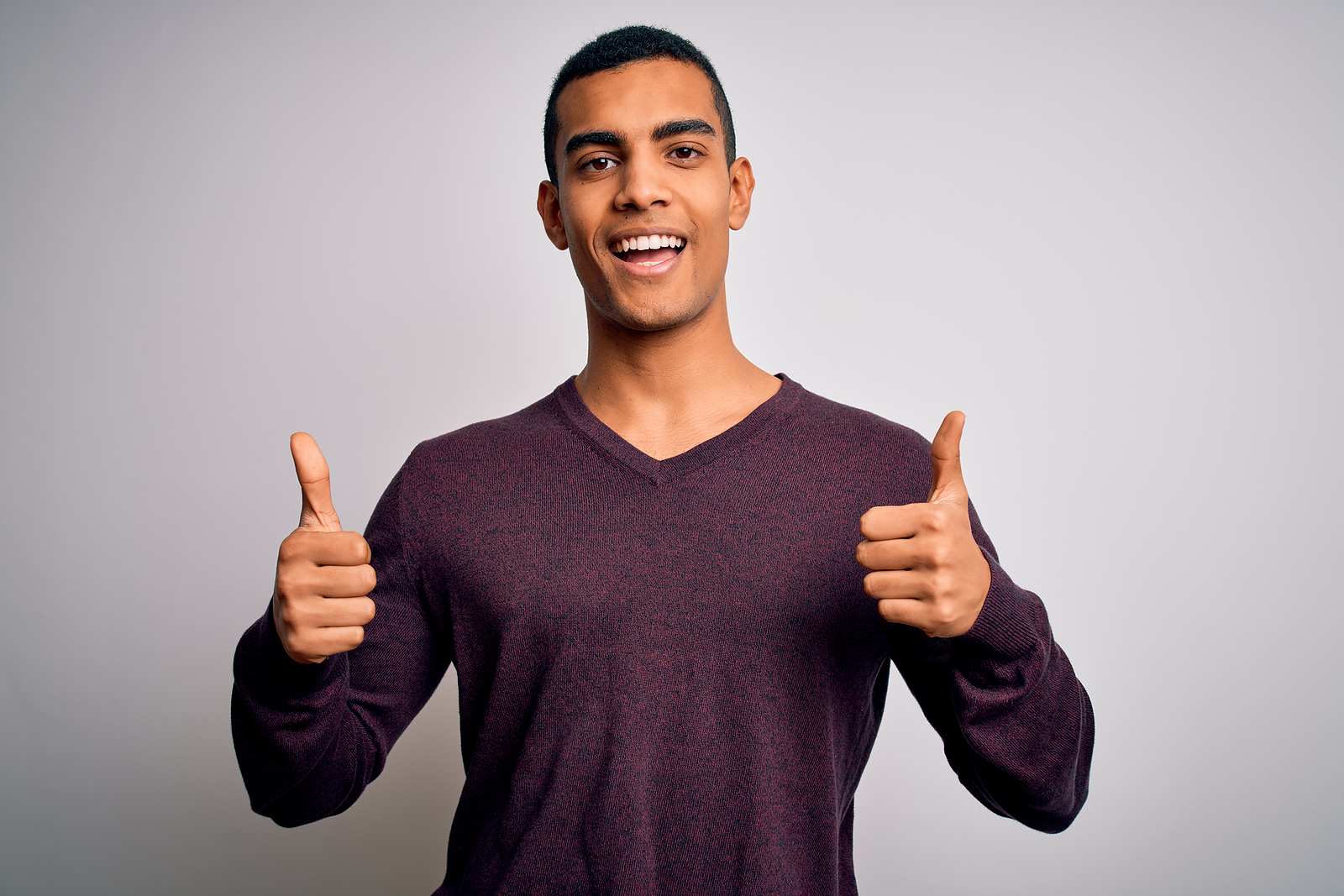 An attractive man smiles and holds his thumbs out in front of a plain background.