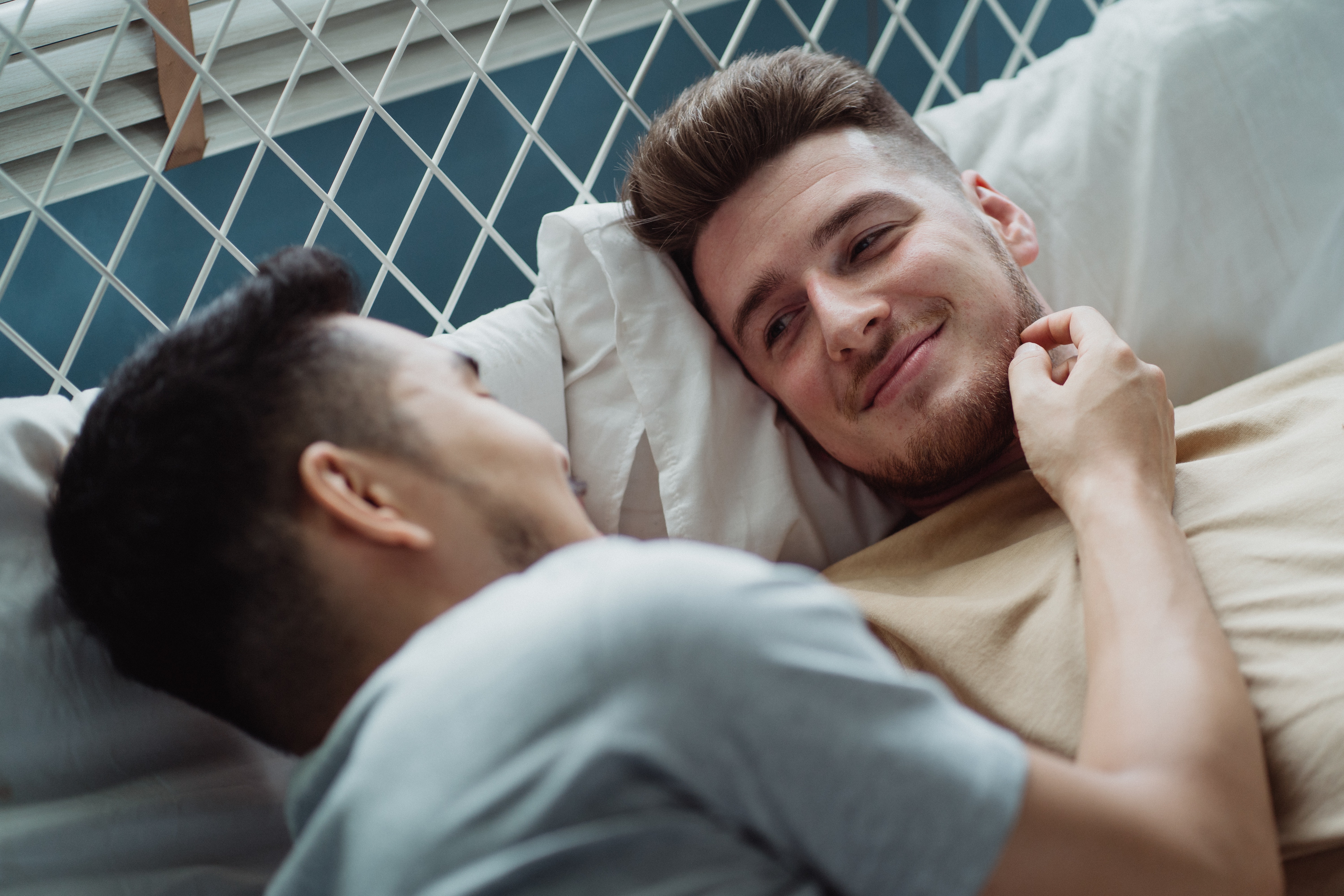 Two young men lying in bed together, one of them has his hand over the others' face and both are smiling.