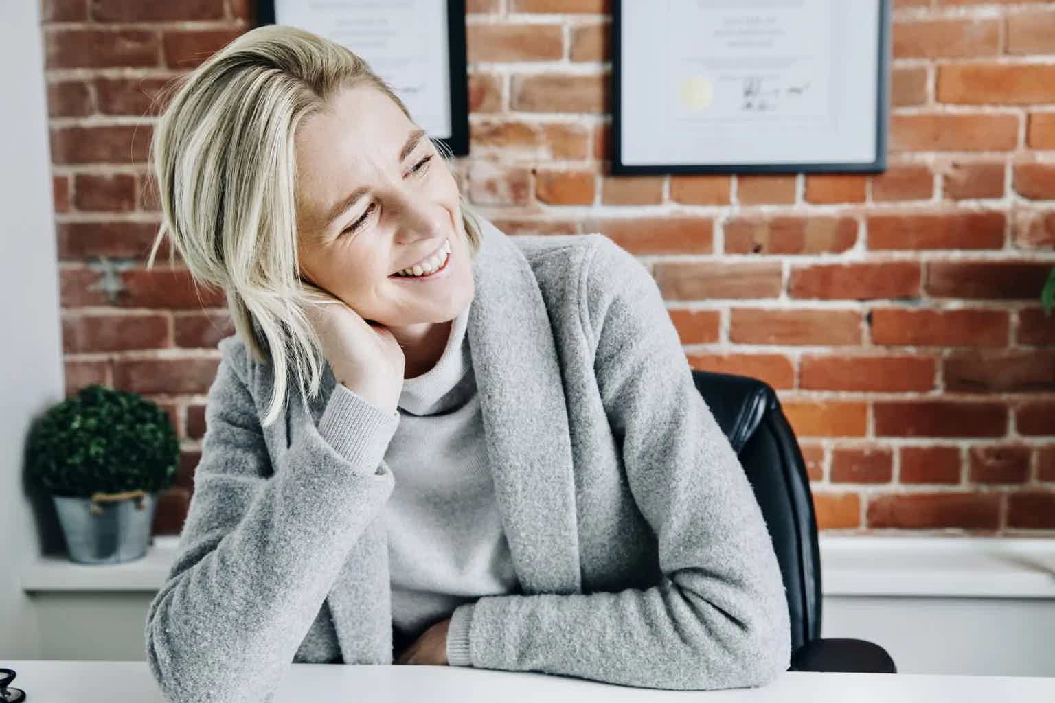 Smiling woman with blonde hair sits at desk, head resting on hand, brick wall and framed certificates in background.
