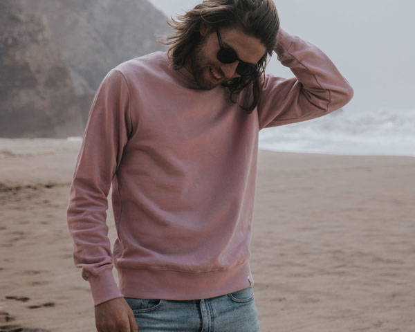 Man wearing soft pink organic cotton sweatshirt on the beach, from sustainable clothing brand Goose Studios, based in Brighton in the UK
