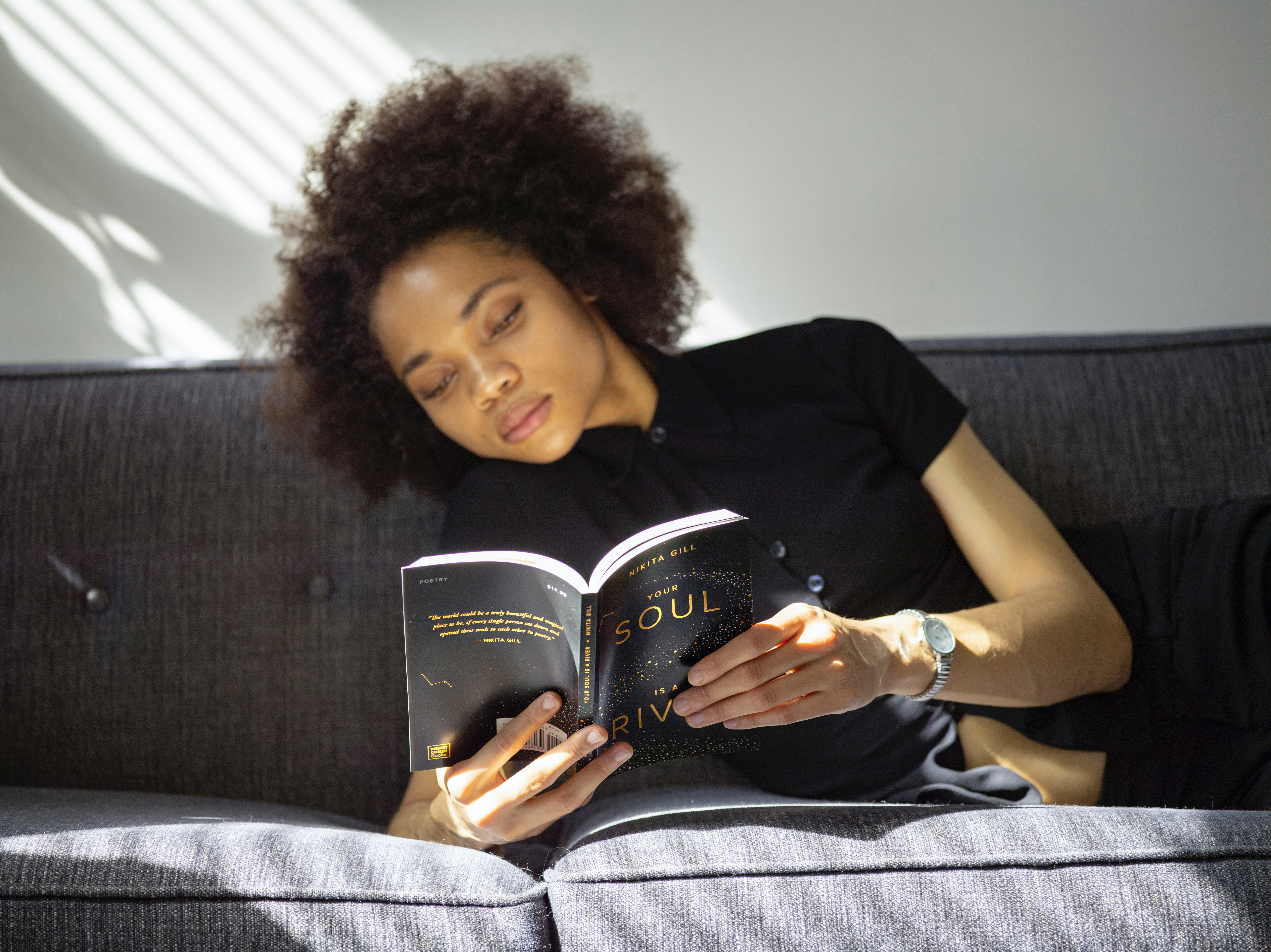 An attractive black woman with large curls leans on a couch and reads a book comfortably.