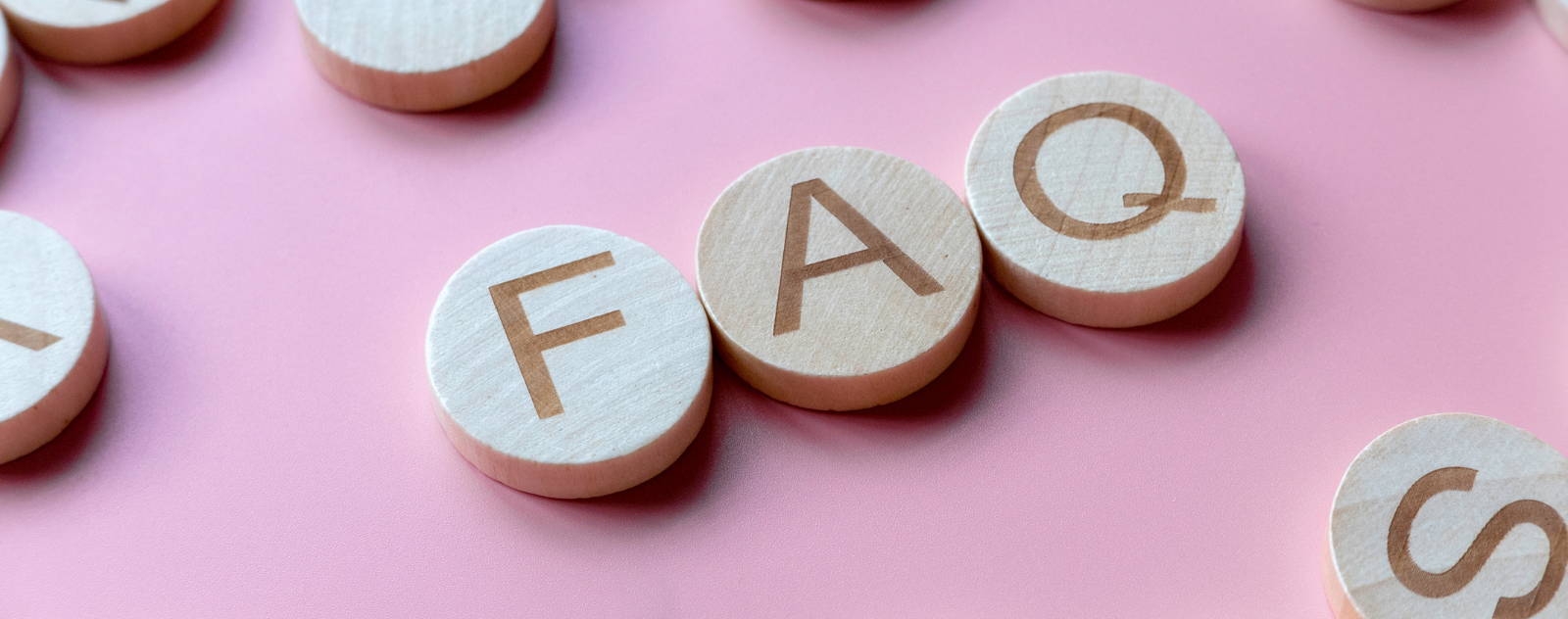 Pièces de jeu rondes en bois avec les lettres 'FAQ' sur un fond rose, représentant des questions fréquemment posées.