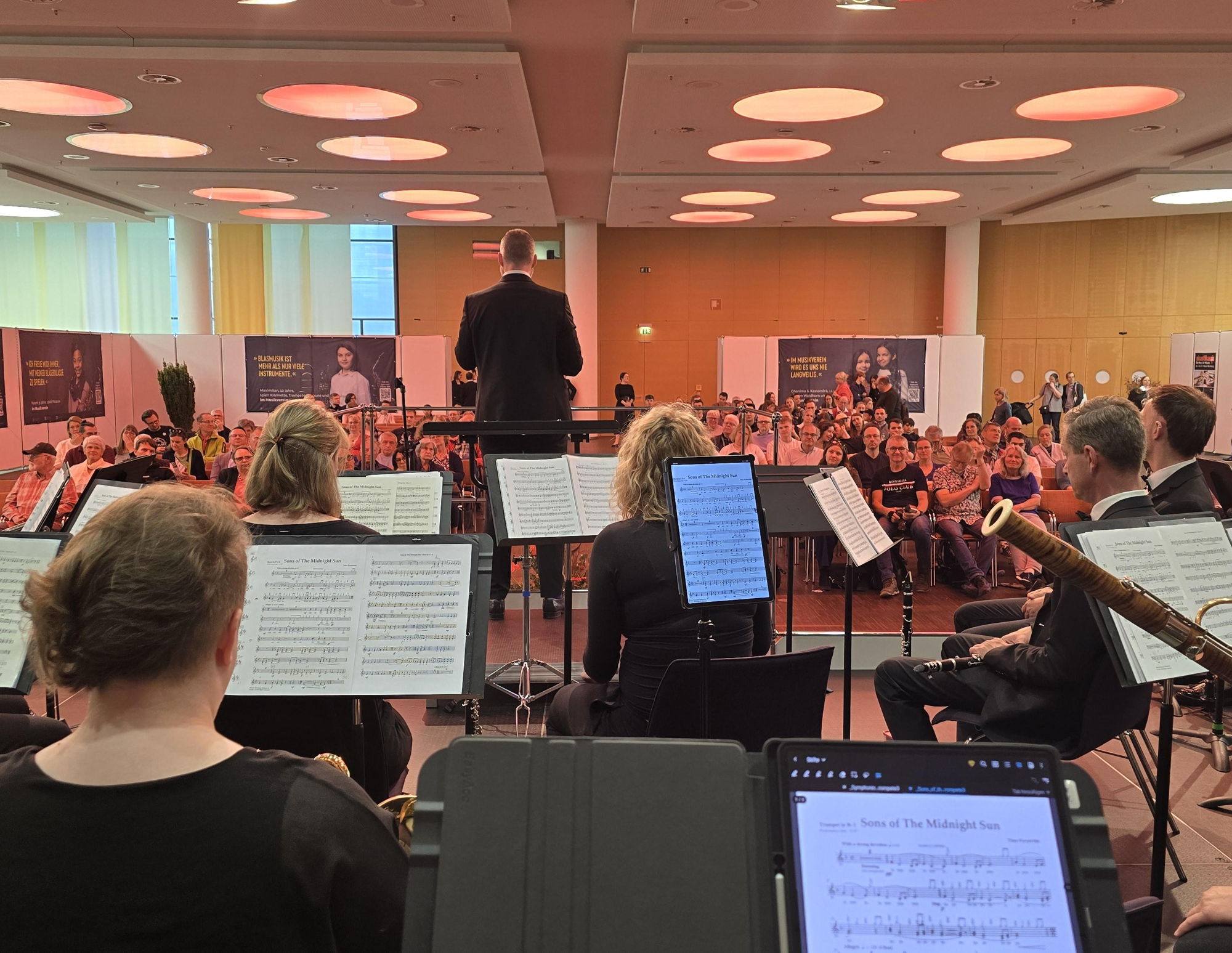 Ein Orchester sitzt auf einer Bühne und musiziert vor großem Publikum. 