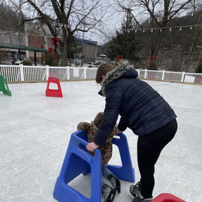 Two individuals ice skating, one assisting the other with a blue aid, evoking joy and camaraderie on a festive winter day.
