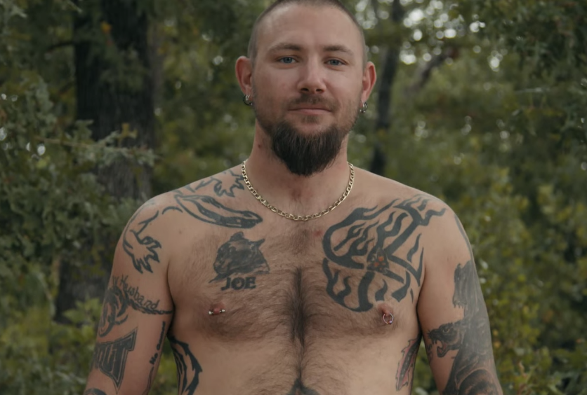 John Finlay smirking while shirtless outdoors. His body has several tattoos and piercings.