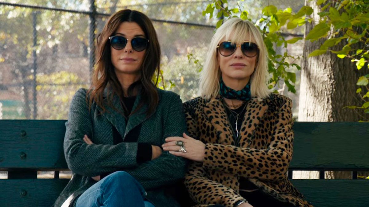 Lou y Debbie sitting side by side wearing sunglasses, their hands touching and smirks on their faces.