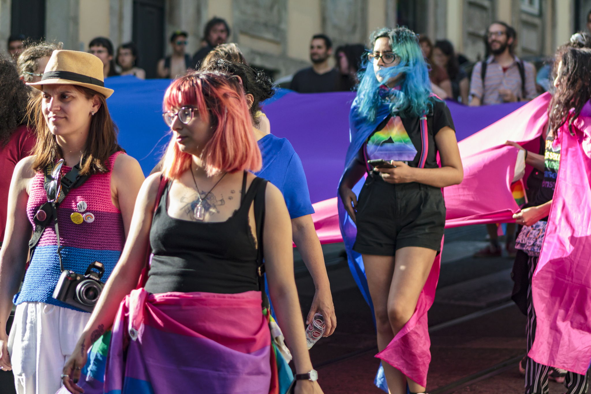 Several women with different colored hair, marching together wearing the bi flag on their bodies.