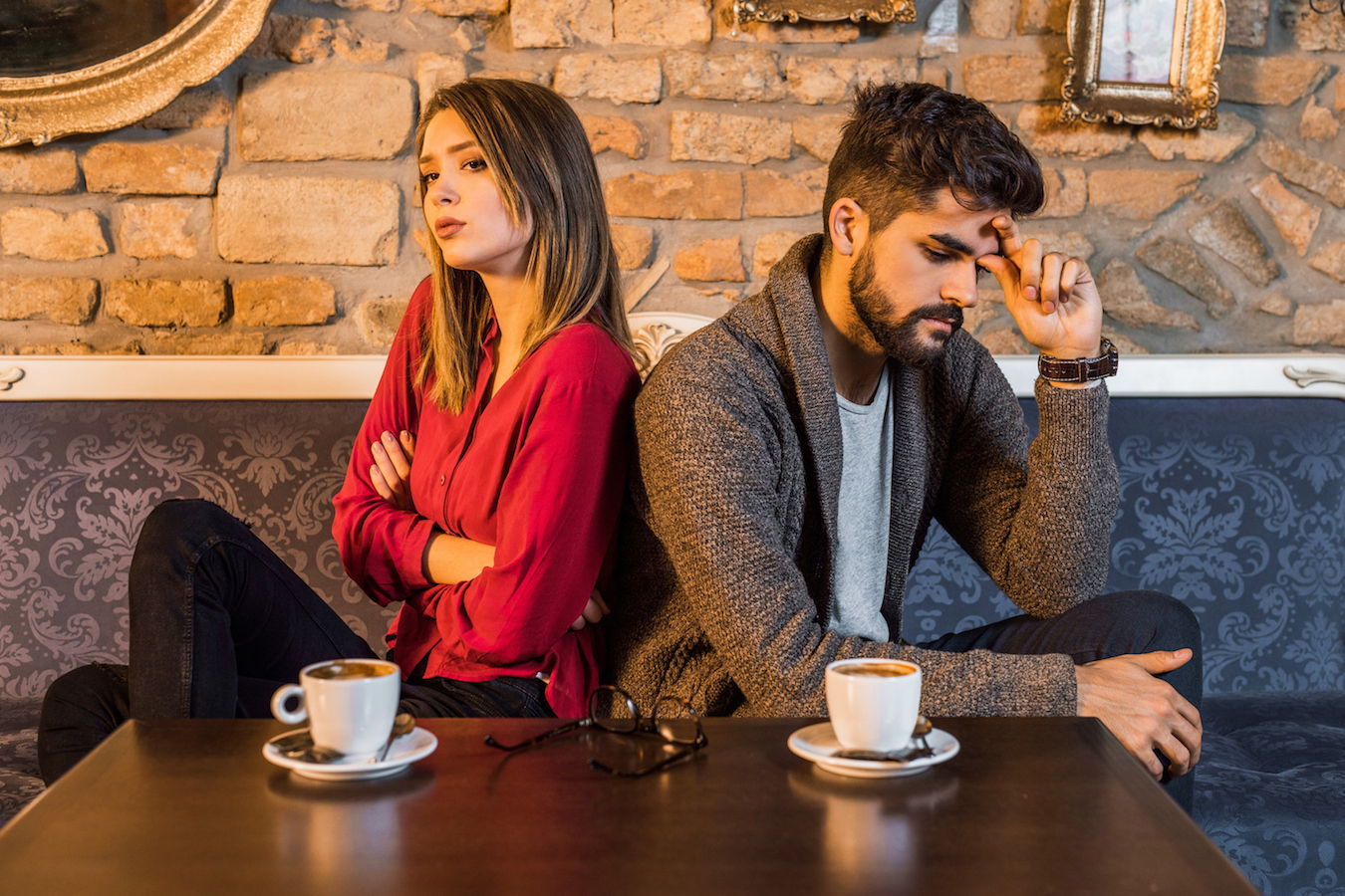 A couple sits back to back at a cafe with annoyed looks on both their faces.