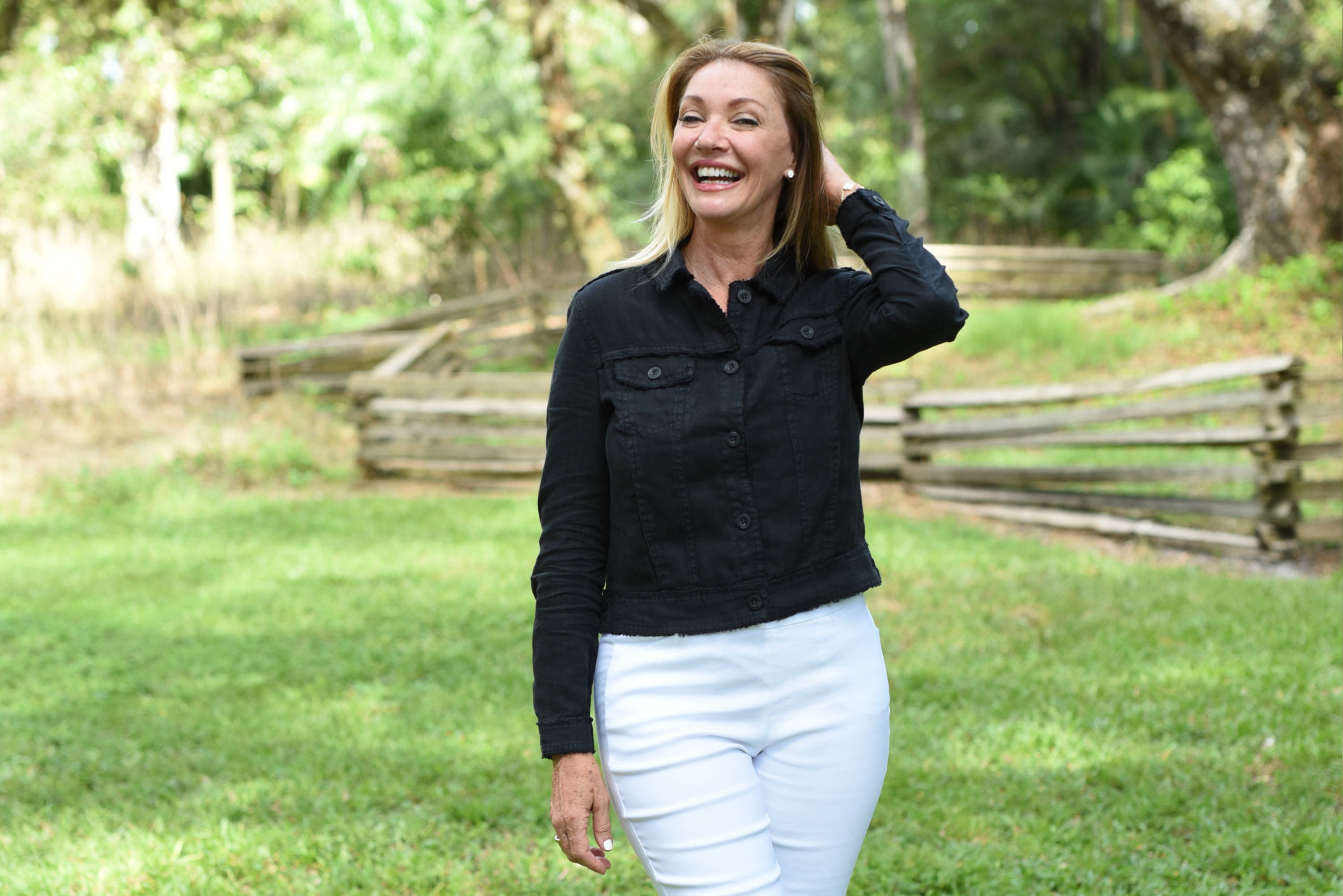 A woman in a black jacket with white denim ankle pants in a sunny green field.