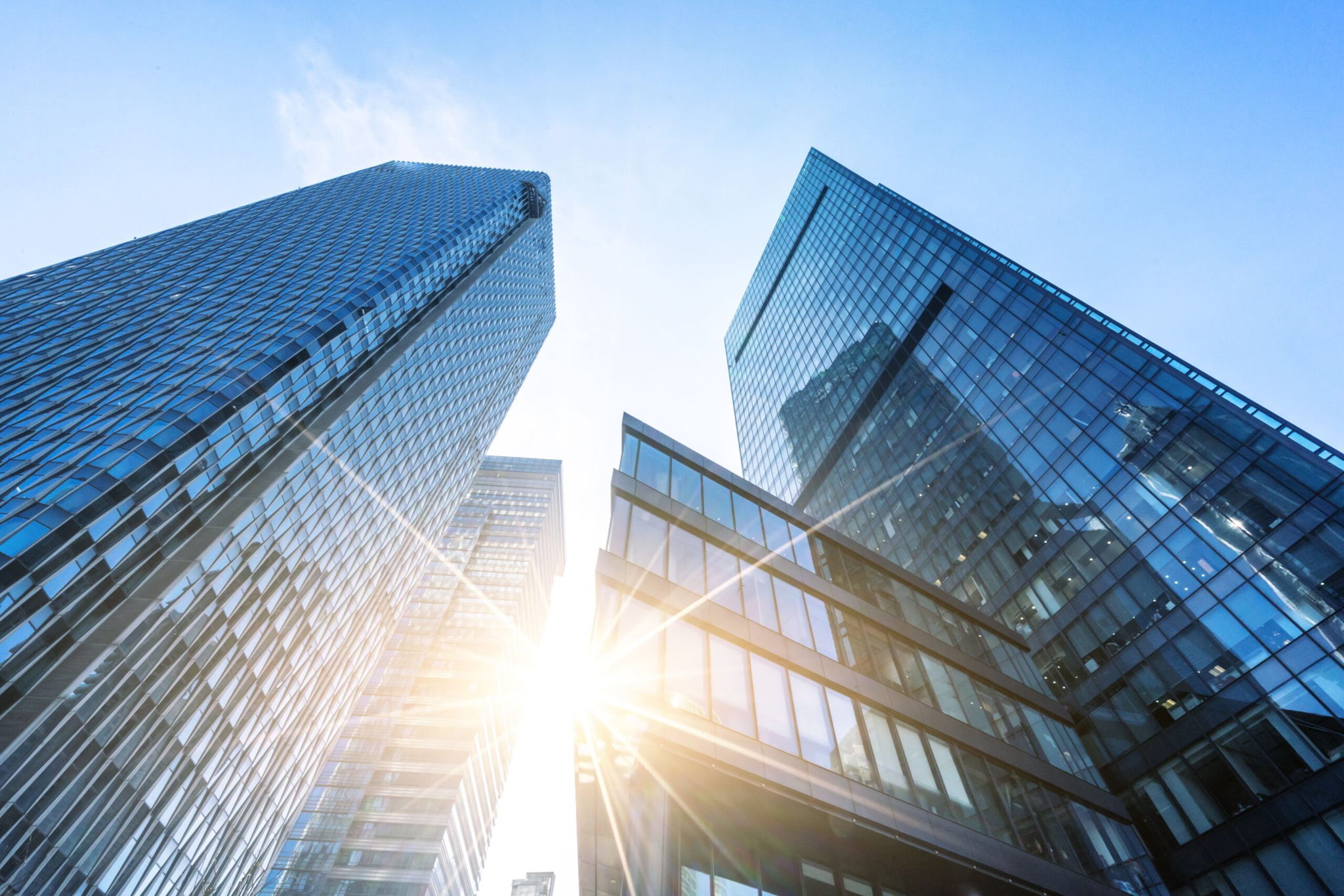 Looking up at several modern glass skyscrapers with the sun shining brightly between them against a clear blue sky. The buildings reflective surfaces create a dynamic play of light and shadow.