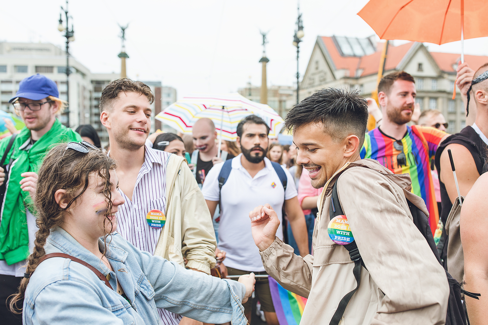 A young man and woman with pride pins on their clothes, dancing in the street while others are walking around them with pride pins as well.