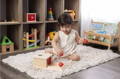 Little girl sitting on a carpet in her playroom and placing red balls into the Object Permanence box, with beautiful Montessori toys on the shelf in the background.