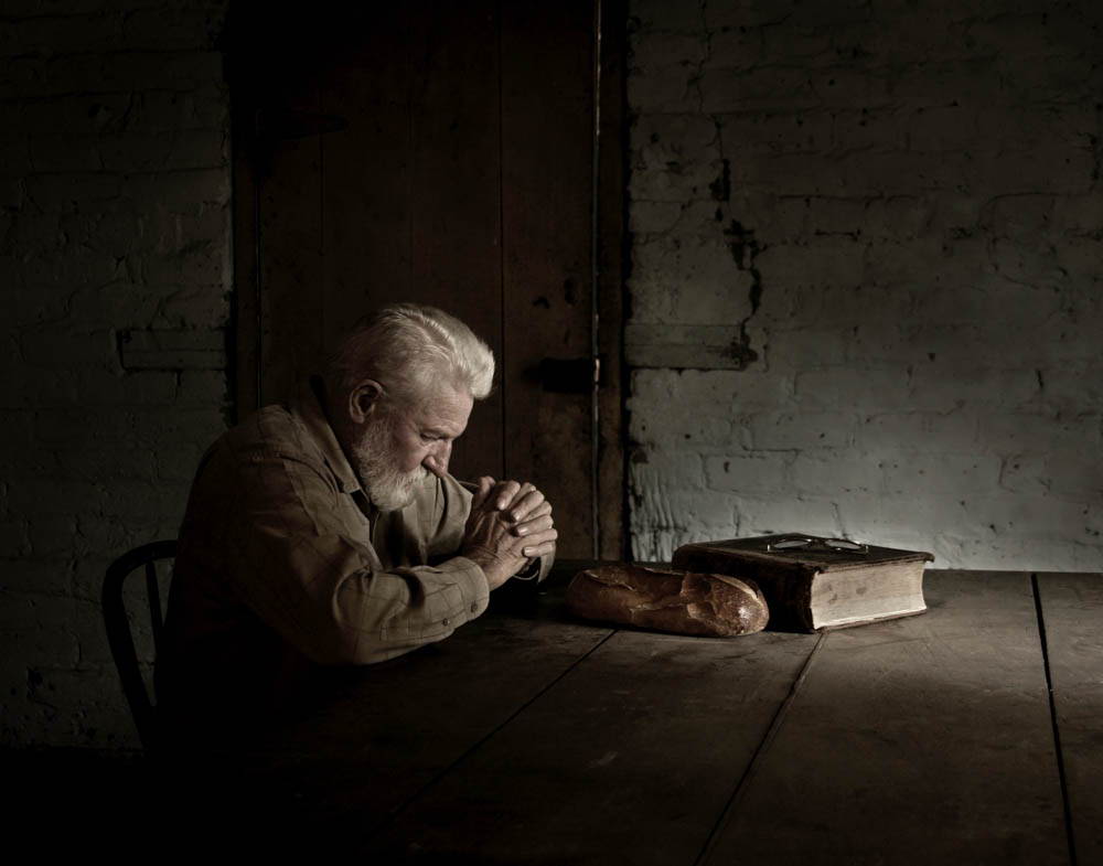 An old, pioneer man praying at his table next to the Bible.