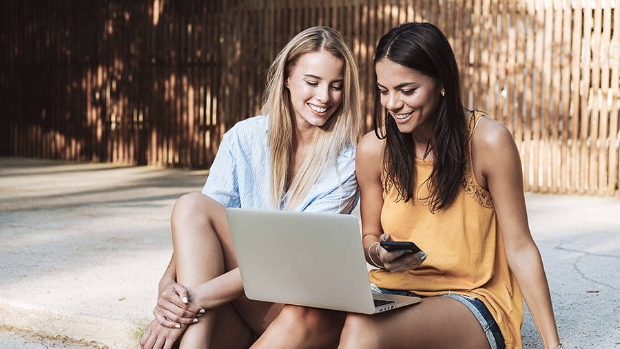 Two smiling friends sitting on a step reading about how to spend reward points. One woman is holding a phone ready to sign up for rewards