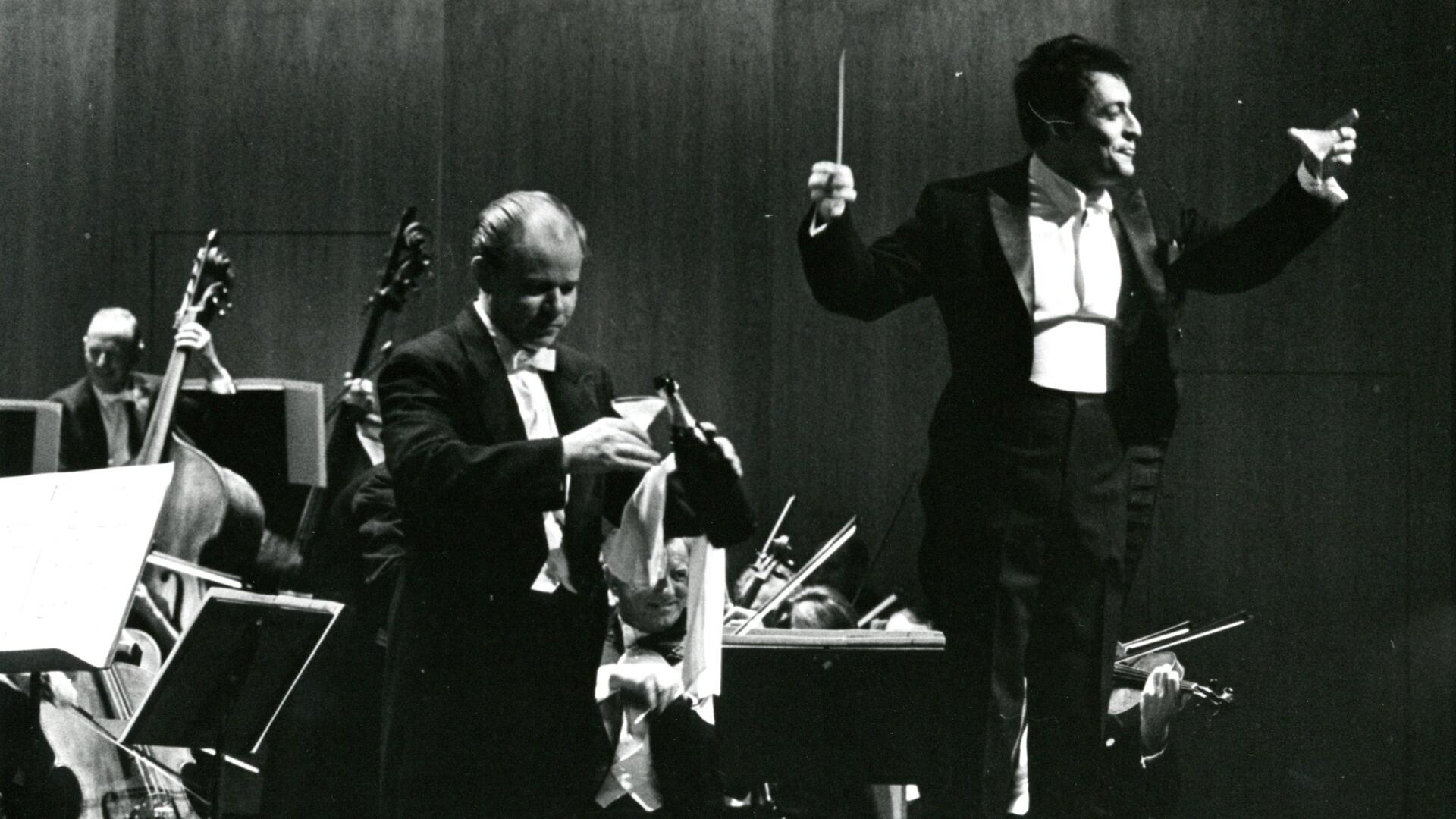 Zubin Mehta stands at the podium in front of the orchestra, raising a glass of champagne to the audience.