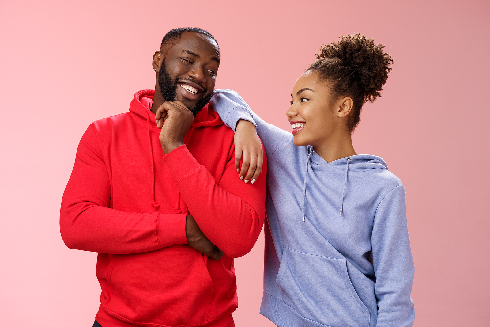 An attractive man and woman smile to eachother. The woman has her arm over his shoulder while his arms are crossed.