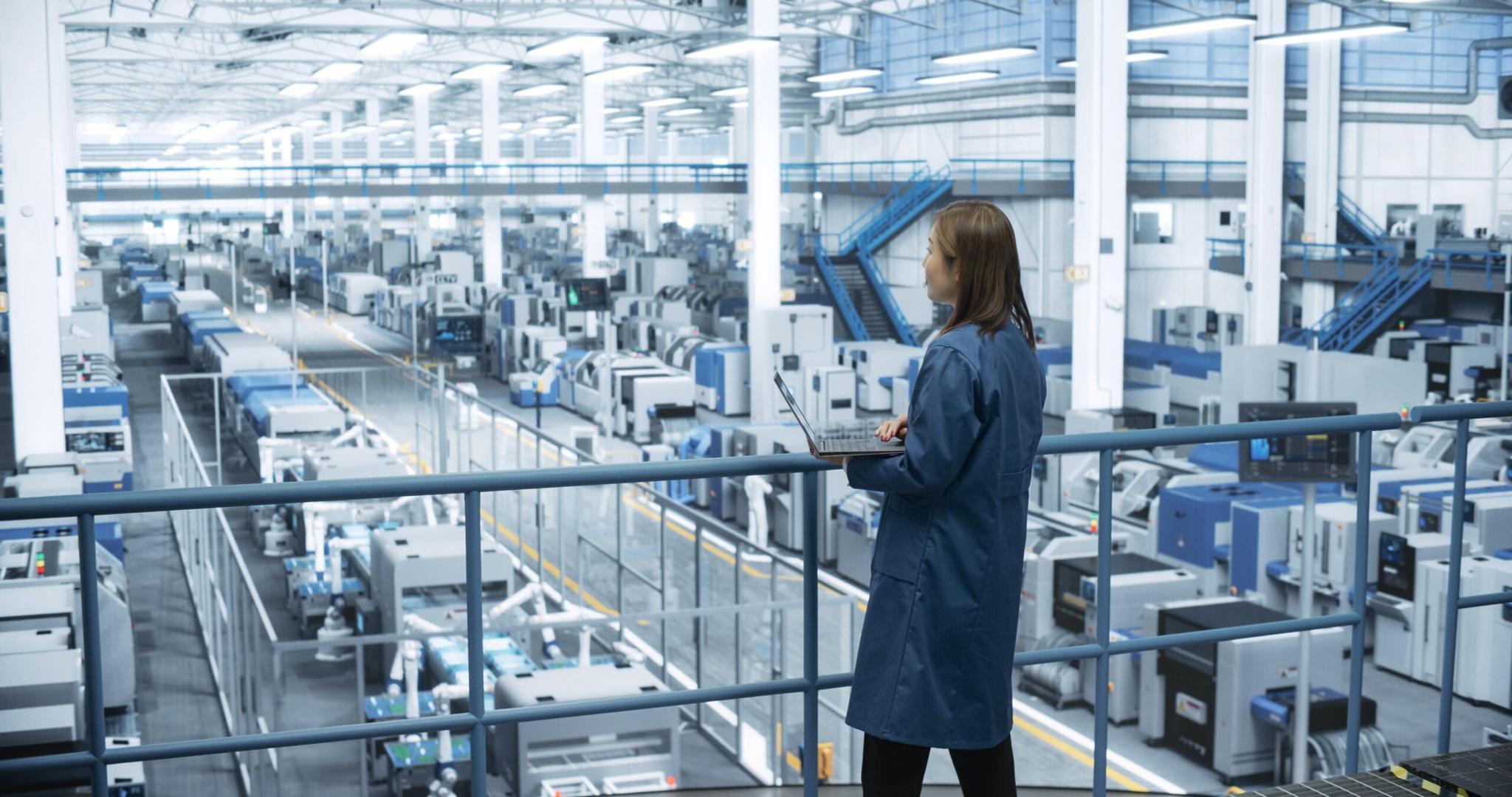 A woman in a blue lab coat stands on a balcony overlooking a large, modern factory floor filled with industrial machines and equipment under bright lighting.