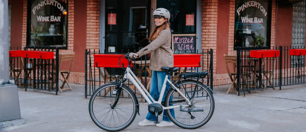 a woman walking next to her electric city bike in front of a shop in a city