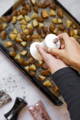 Diced potatoes lay on a baking sheet beneath a set of hands using a white grinding mill