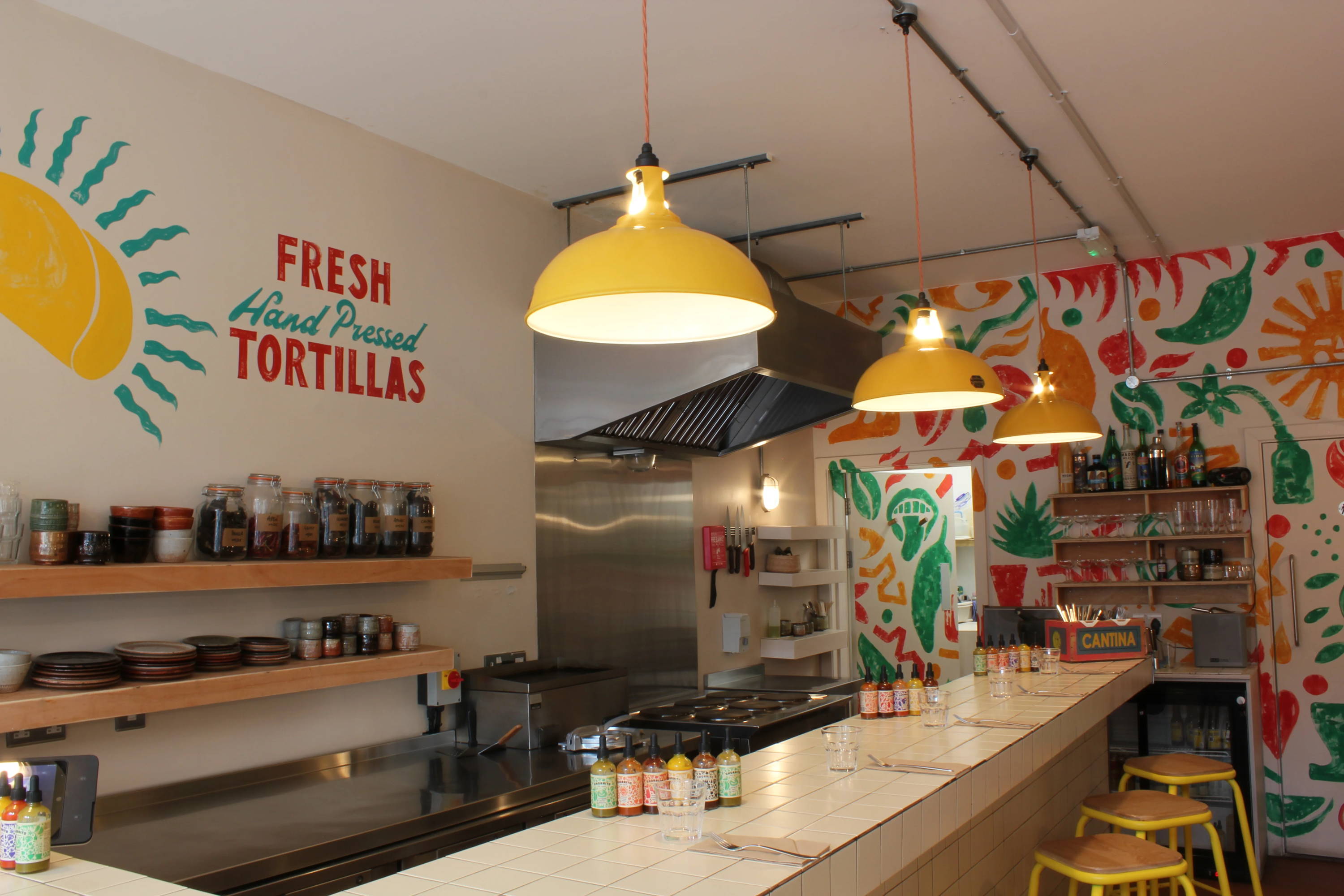 Three Large Yellow Pendant Lampshades hang in a row over a tiled bar, separating the kitchen area behind it, with Yellow barstools on the other side. On the back walls is hand-painted artwork in bright colours.
