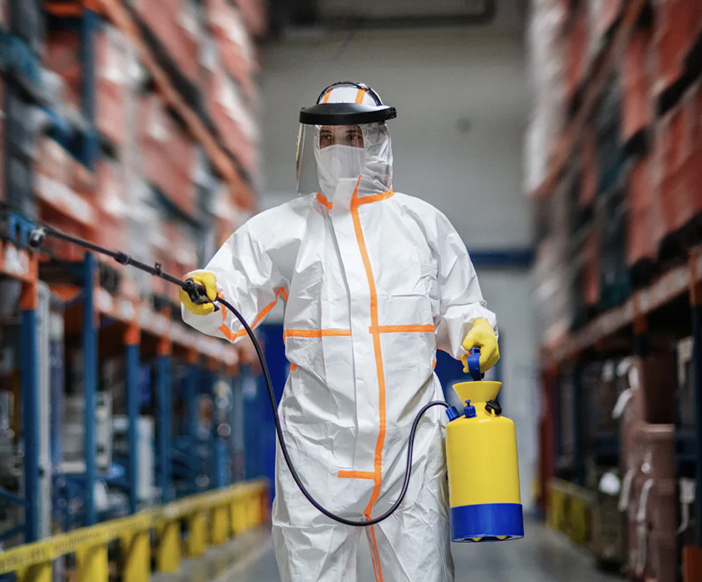 A person in full protective gear, including a face shield, gloves, and a white hazmat suit, sprays disinfectant in a warehouse aisle with shelves stacked with boxes.