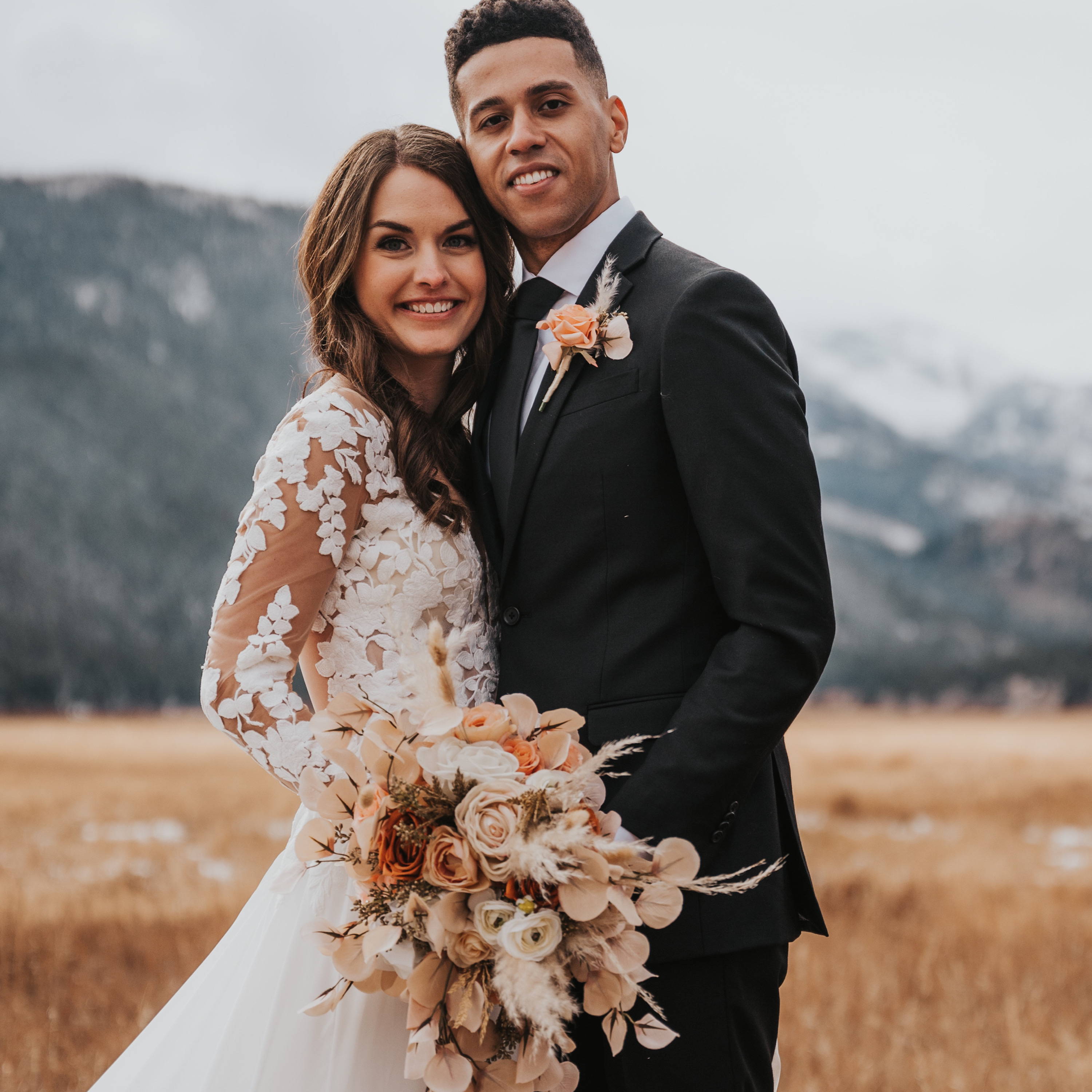 A newly married couple, the bride is holding a cascading bridal bouquet that is very boho inspired. It is beige, rust, and orange with pampas grass and lots of roses. 