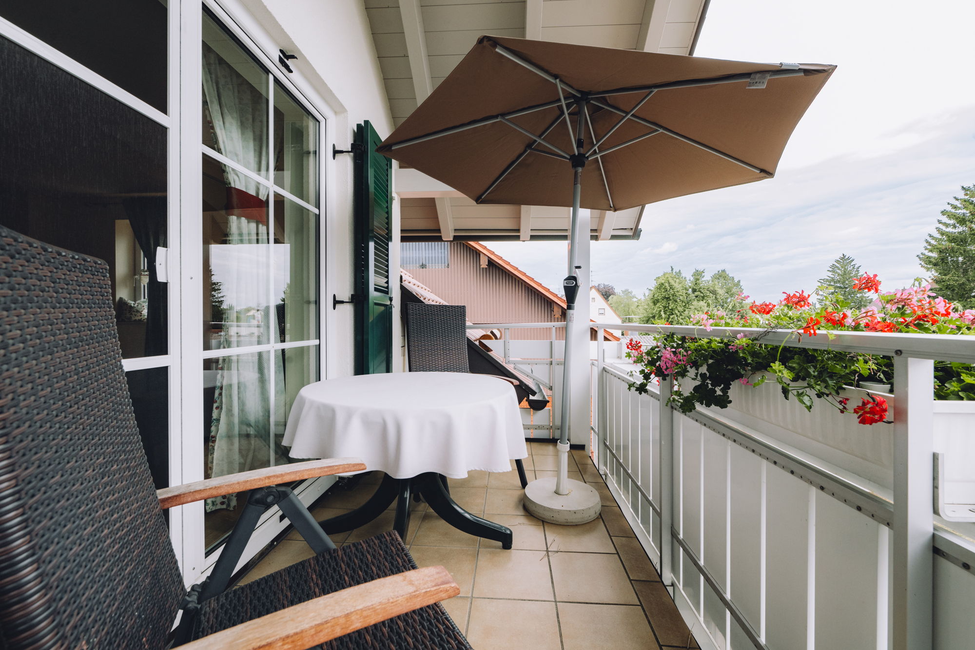 Ein Balkon mit Sonnenschirm, weißem Tisch, verstellbaren Stühlen und Blumenkasten.
